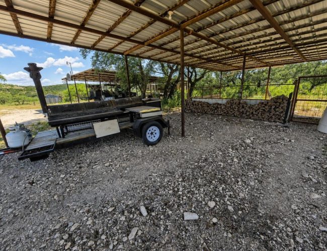 3E Ranch smoker trailer and stacked firewood under a metal roof